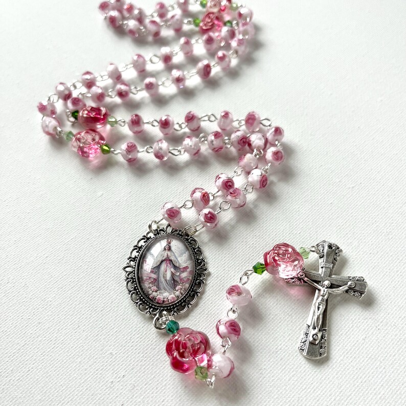 May include: A rosary featuring pink and white rose-shaped beads connected by silver-toned metal links. The piece includes a silver-toned crucifix and an oval-shaped centerpiece medallion depicting a figure in a robe, accented with small green beads.
