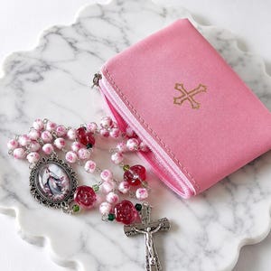 May include: A pink zippered pouch featuring a gold cross emblem on the front, accompanied by a rosary with pink rose-patterned beads, translucent red rose-shaped beads, a silver-toned crucifix, and an oval pendant depicting a religious figure.
