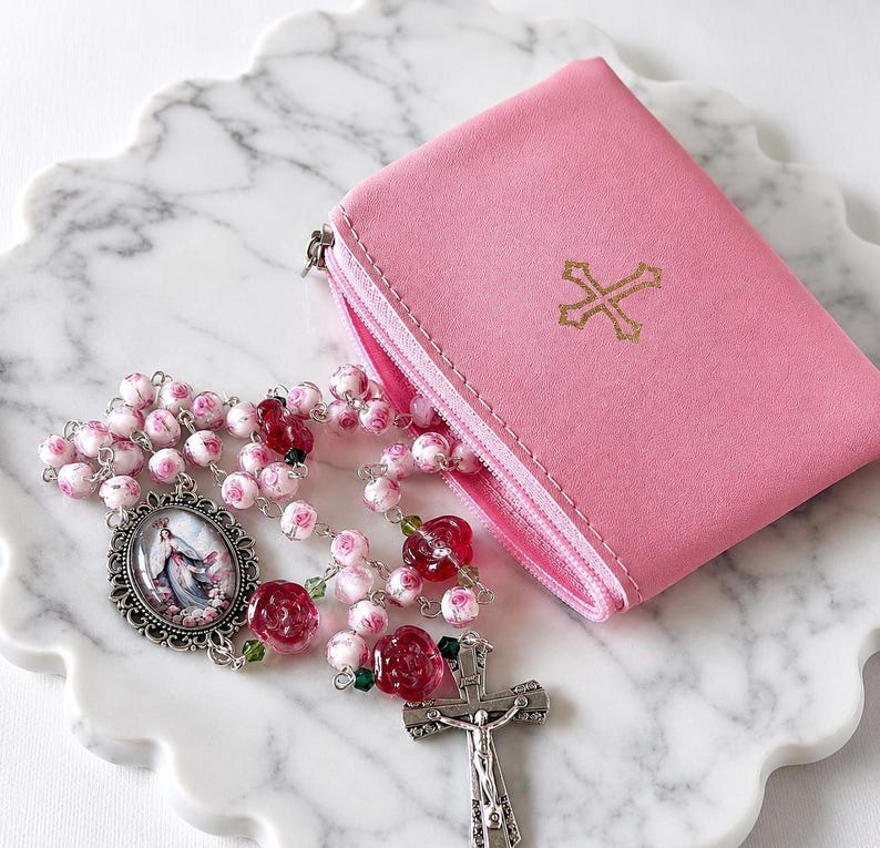 May include: A pink zippered pouch featuring a gold cross emblem on the front, accompanied by a rosary with pink rose-patterned beads, translucent red rose-shaped beads, a silver-toned crucifix, and an oval pendant depicting a religious figure.