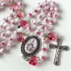 May include: A rosary featuring pink and white rose-shaped glass beads linked by silver-toned metal chain. The centerpiece is an oval pendant depicting a figure in a gown, and the crucifix is a silver-toned metal cross with a detailed figure.