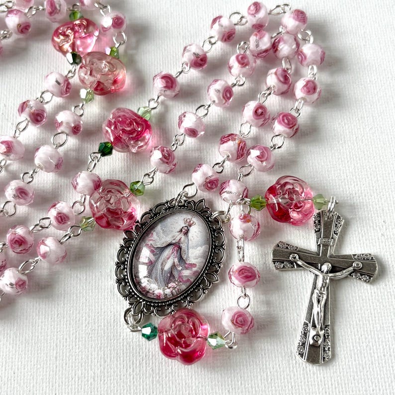 May include: A rosary featuring pink and white rose-shaped glass beads linked by silver-toned metal chain. The centerpiece is an oval pendant depicting a figure in a gown, and the crucifix is a silver-toned metal cross with a detailed figure.