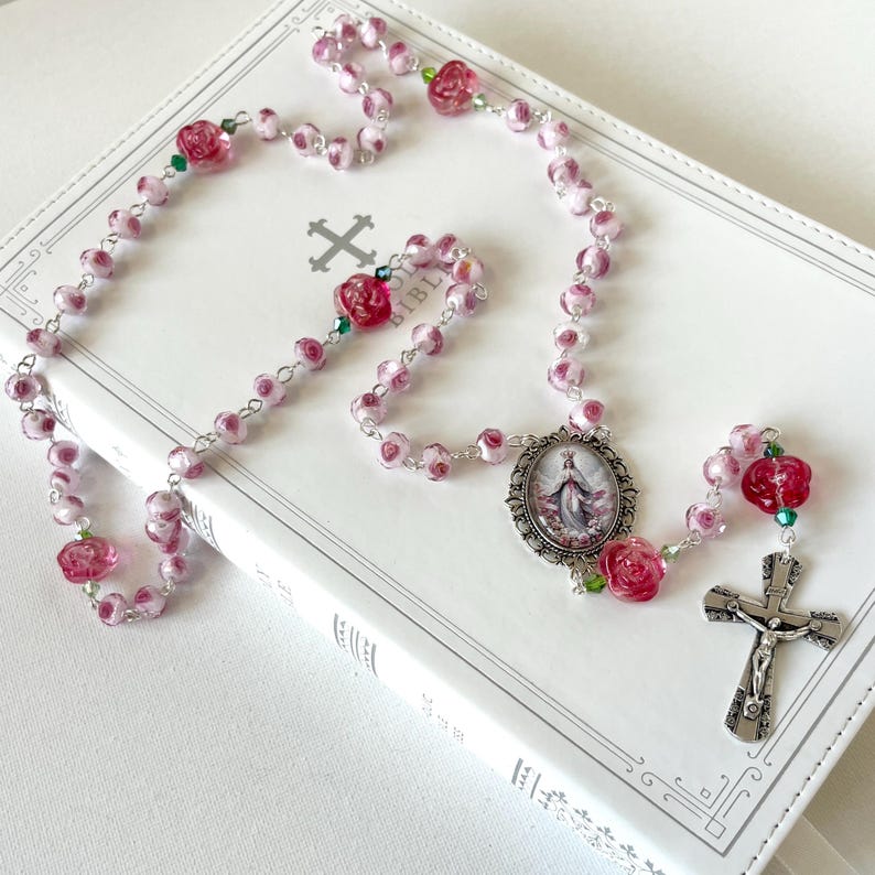 May include: A rosary featuring pink and white marbled beads interspersed with larger, deep pink rose-shaped beads and small green crystals. The rosary includes a silver-toned metal crucifix and an oval centerpiece depicting a religious figure.