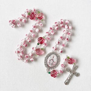 May include: A rosary featuring beads shaped like pink and white roses, interspersed with small green crystal accents. The centerpiece is an oval metal medallion depicting a figure, and the end features a silver-toned metal crucifix.