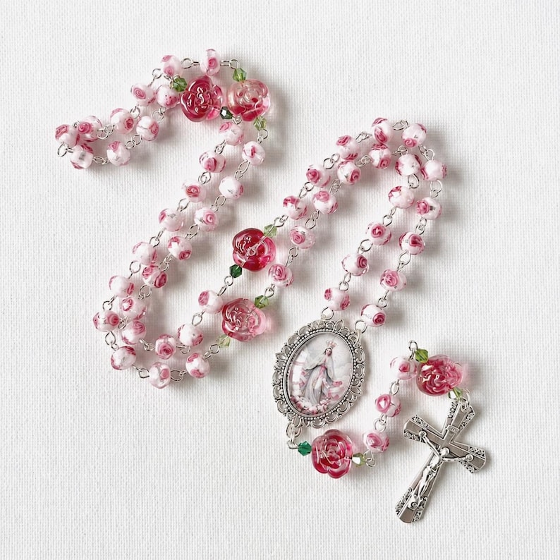 May include: A rosary featuring beads shaped like pink and white roses, interspersed with small green crystal accents. The centerpiece is an oval metal medallion depicting a figure, and the end features a silver-toned metal crucifix.