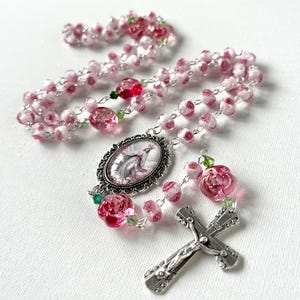 May include: A rosary featuring pink and white rose-shaped glass beads, small green crystal accents, and a silver-toned metal crucifix. An oval centerpiece displays a religious figure in a decorative silver frame, connected by silver-toned metal links.