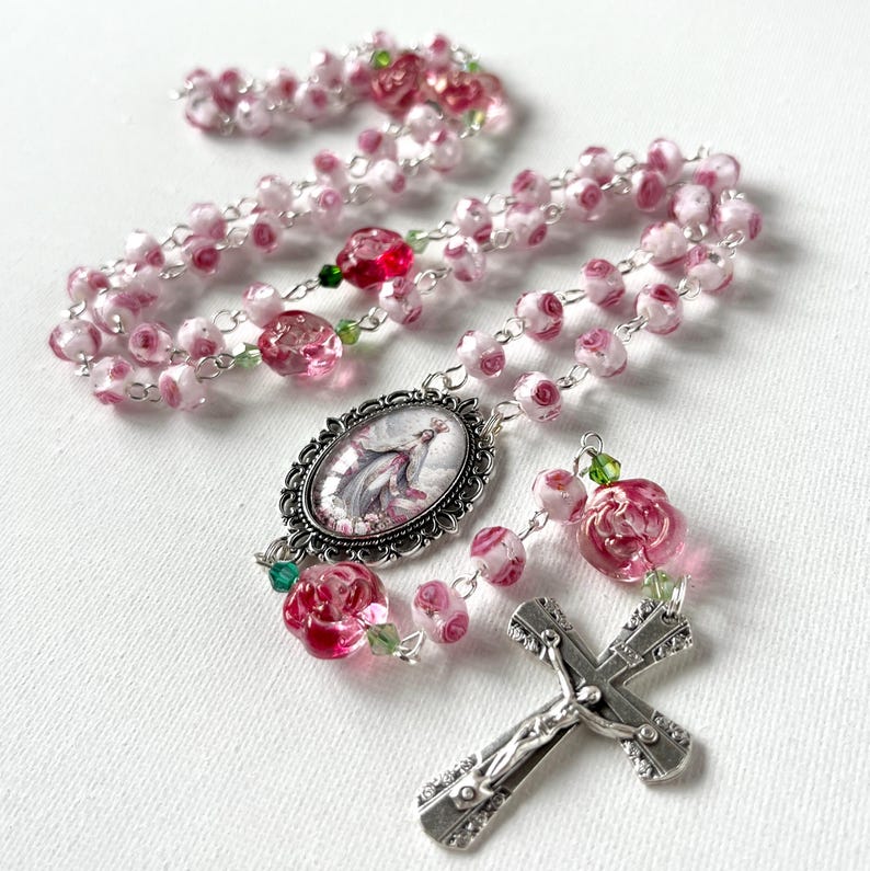 May include: A rosary featuring pink and white rose-shaped glass beads, small green crystal accents, and a silver-toned metal crucifix. An oval centerpiece displays a religious figure in a decorative silver frame, connected by silver-toned metal links.