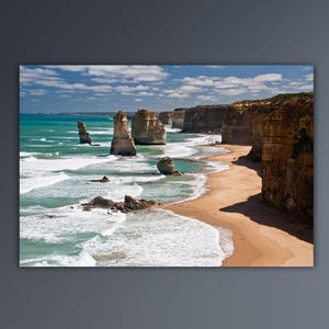Puede incluir: Una vista panorámica de los Doce Apóstoles, un conjunto de pilas de piedra caliza a lo largo de la costa de Victoria, Australia. Las pilas se elevan desde el océano turquesa, con olas blancas que rompen en la orilla. Una playa de arena se extiende a lo largo de la base de los acantilados, con un acantilado escarpado en el lado derecho de la imagen.