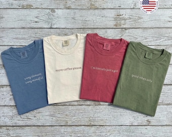 May include: Four short-sleeved t-shirts in blue, cream, red, and olive green are displayed. Each shirt has a different embroidered phrase: "very demure very mindful," "more coffee please," "I'm literally just a girl," and "good vibes only."