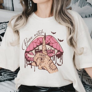 Può includere: T-shirt color crema con una grafica raffigurante una bocca con zanne, un dito con un tatuaggio con la scritta "TRUST" e le parole "Basic witch". Include pipistrelli e dettagli di sangue che cola.