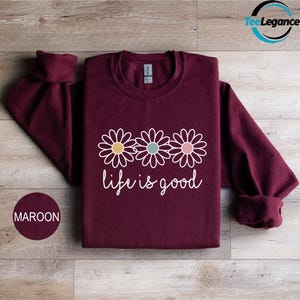 May include: Maroon sweatshirt with three daisy flowers and the words "life is good" in white script. The flowers have yellow, teal, and pink centers. The sweatshirt has long sleeves.