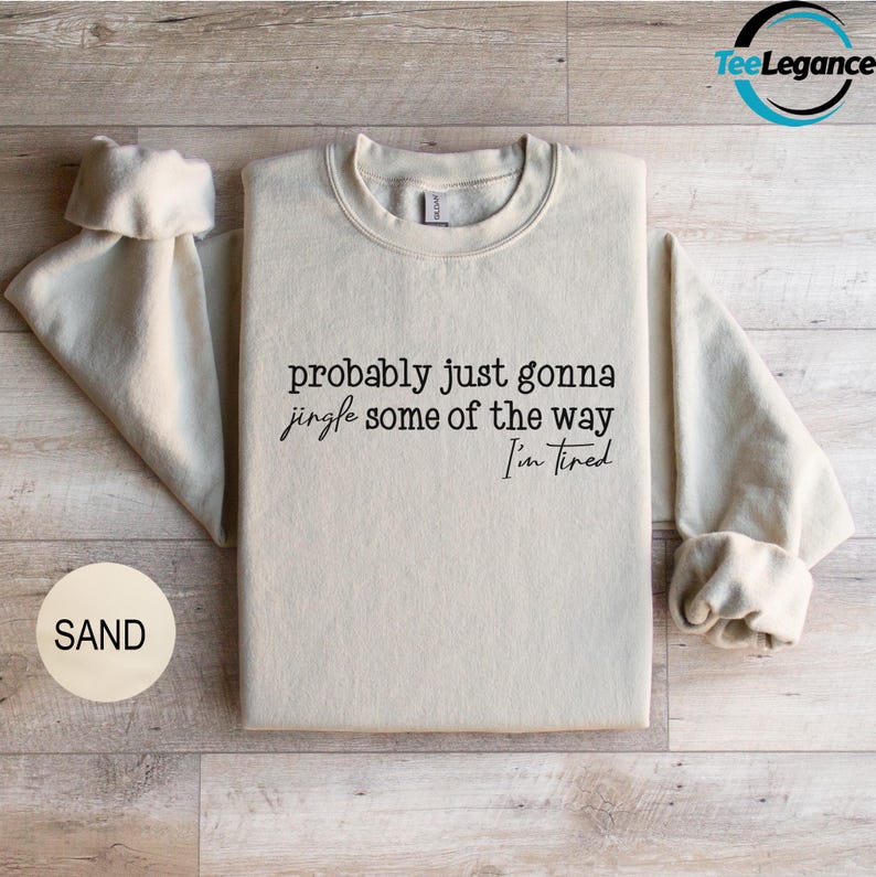 A beige t-shirt with the text "probably just gonna jingle some of the way I'm tired" printed on it, along with a small circular logo that says "SAND".