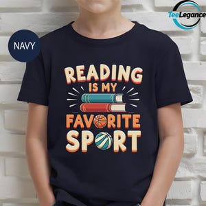 May include: Navy blue t-shirt with the text "READING IS MY FAVORITE SPORT." The design features stacked books, a basketball, and a volleyball. The text and graphics are in shades of orange, white, blue, and red.