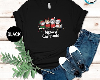 May include: Black t-shirt featuring a festive design with four cartoon cats dressed in Christmas attire. The cats are adorned with elf hats, reindeer antlers, and Santa hats, with the words "Meowy Christmas" below.