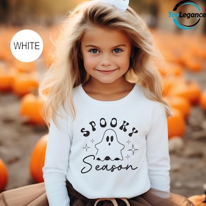 May include: A young girl wearing a white long-sleeved shirt with the words "Spooky Season" and a ghost graphic. The shirt is paired with a brown skirt. The background features a pumpkin patch.