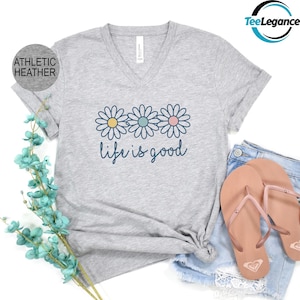 May include: Athletic heather gray V-neck t-shirt with a daisy graphic and the text "life is good" in blue. The shirt is paired with denim shorts and pink flip-flops. The shirt is made of a soft, comfortable material.