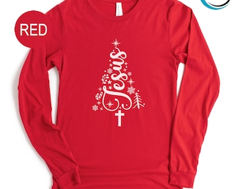 May include: A red long-sleeve shirt with a white Christmas tree design. The tree is made up of the word "Jesus", snowflakes, stars, and a cross. The word "RED" is in a red circle in the upper left corner.