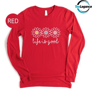 May include: A red long-sleeve shirt with a white floral design and the phrase "life is good" in cursive. The flowers have yellow, blue, and pink centers. The shirt has a crew neck.