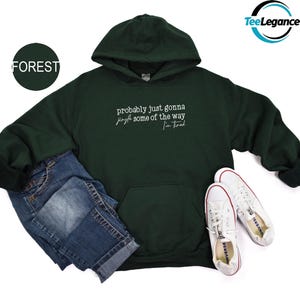 A green hoodie with white text, a pair of blue jeans, and white sneakers are arranged on a white background.