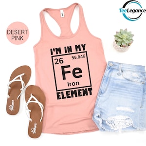 May include: A desert pink tank top with the text "I'M IN MY Fe Iron ELEMENT" in black. The image also includes denim shorts, brown sandals, and a small potted succulent.