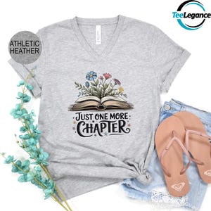 May include: Gray V-neck t-shirt with a graphic of an open book with flowers growing out of it. The text "Just One More Chapter" is written below the book.