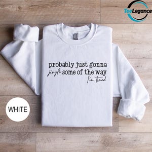 A white t-shirt with the text "probably just gonna jingle some of the way I'm tired" printed on it, laid out on a wooden surface.