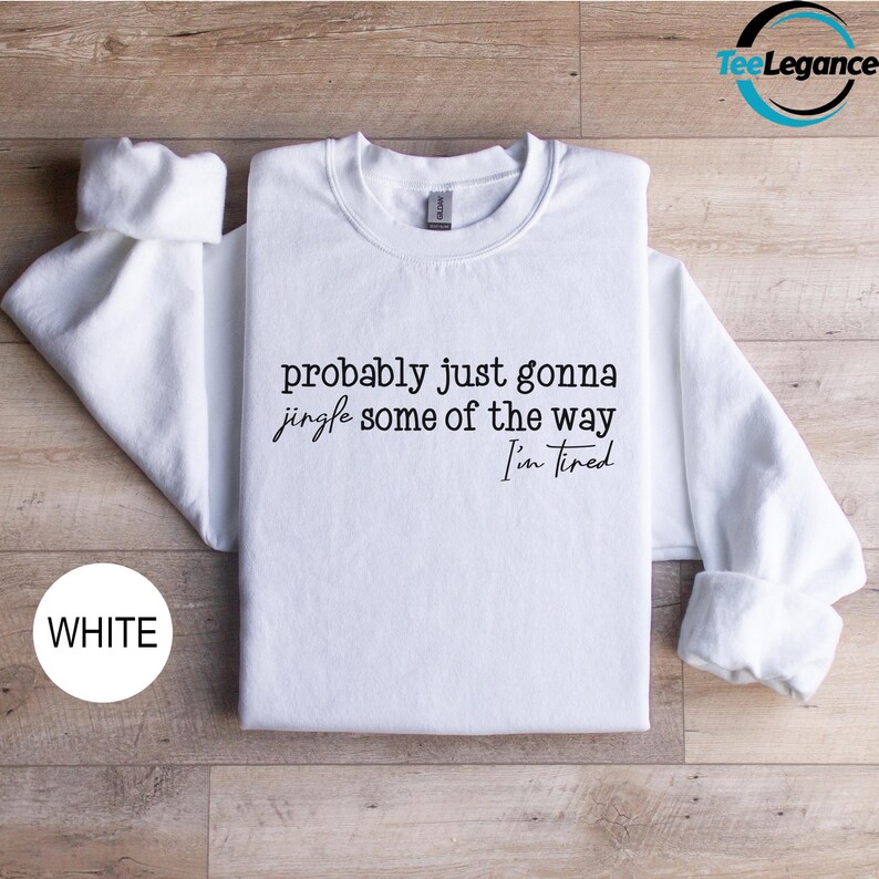 A white t-shirt with the text "probably just gonna jingle some of the way I'm tired" printed on it, laid out on a wooden surface.