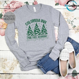 May include: Athletic heather long-sleeve shirt with a green graphic of pine trees and the text "Columbia Inn Pine Tree, Vermont America's Snow Playground". The shirt is paired with blue jeans and white flats.