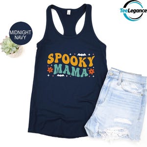 May include: Midnight navy tank top with the words "SPOOKY MAMA" in orange, teal, and white. The design includes bats, flowers, and stars. Light blue denim shorts are next to the tank top.