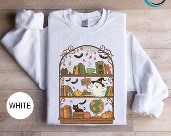 May include: White sweatshirt featuring a Halloween-themed bookshelf design. The graphic includes books, pumpkins, a ghost holding a "BOO-KS" sign, a black cat, bats, and a globe. The design is in shades of orange, green, brown, and black.