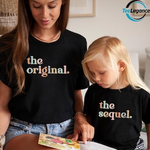May include: Two black t-shirts with the phrases "the original." and "the sequel." printed in pastel colors. The shirts are worn by a woman and a child, who are looking at a book together. The image has a warm, family-oriented theme.
