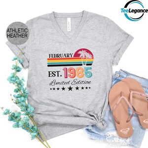 May include: A light gray V-neck t-shirt with a colorful graphic design. The design includes the text "FEBRUARY EST. 1985 Limited Edition" with a sunset and palm tree illustration. The shirt is paired with denim shorts and sandals.