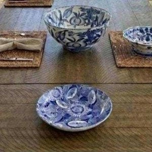 May include: A large round bowl, a smaller scalloped bowl, and a flat plate, all featuring a blue and white floral pattern. The set is arranged on a wooden surface alongside a folded beige napkin and metal cutlery placed on woven rectangular mats.