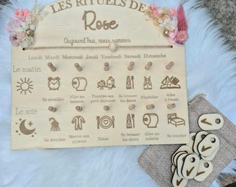 May include: Wooden daily routine chart with the text "LES RITUELS DE Rose" and "Aujourd'hui, nous sommes". The chart has pegs for each day of the week and icons representing daily activities. Includes wooden tokens.