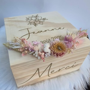 Peut inclure: Un coffret en bois clair avec un motif floral et le nom "Jeanne" gravé sur le couvercle. La boîte est ornée d'une composition florale de fleurs séchées dans les tons de rose, blanc et jaune. Le mot "Merci" est également gravé sur la boîte.