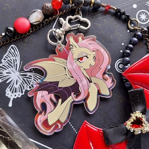 May include: A pink and yellow cartoon character with bat wings and a red and black background. The character has a pink mane and tail and is wearing a black dress. The character is a vampire.