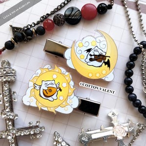 Puede incluir: Dos pinzas para el pelo de metal con diseños de gatos de dibujos animados. Una pinza presenta un gato tocando un laúd dentro de un sol amarillo con nubes y estrellas. La otra pinza muestra un gato tocando el violín en una luna creciente amarilla con estrellas y nubes.