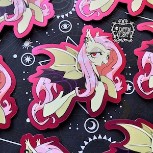 May include: A pink and yellow cartoon character with bat wings and a purple cape. The character has a pink mane and a yellow body. The character is on a red background with black stars and a white crescent moon.
