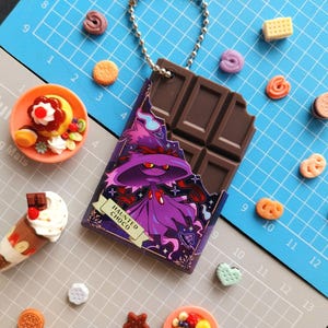 May include: A chocolate bar-shaped keychain with a purple witch design and the text "HAUNTED CHOCO". Miniature desserts and cookies, including a sundae, are scattered around the keychain on a blue and gray cutting mat.