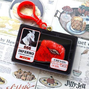 May include: A black and white acrylic keychain featuring a red and white illustration of a mouth with teeth. The keychain also features a black and white illustration of a goat with the text "666 INFERNO CORPORATION LTD." and three stars. The keychain is attached to a red plastic ring.