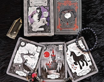 May include: A set of tarot cards with a gothic aesthetic. The cards feature illustrations of a tree, a castle, and a rider. The cards are part of a deck called "Creepy Cat Lenormand" with 40 cards.