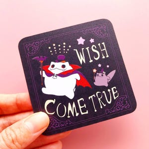May include: A square sticker with a dark purple background featuring a white creature in a red-lined cape and purple top hat holding a staff, alongside a small purple creature with a wand, with the text WISH COME TRUE in white lettering.