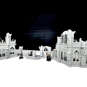 May include: A miniature, gray, ruined gothic building set. The set includes multiple building sections with arched windows and battlements. Small figurines are placed within the structures, suggesting a tabletop game setting.