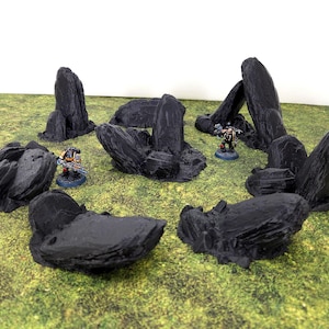 May include: A set of black, textured rock formations on a green surface. Miniature figures are placed amongst the rocks, suggesting a tabletop gaming environment. The rocks vary in size and shape, creating a landscape.