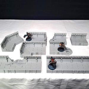 May include: A set of grey, modular terrain pieces for tabletop gaming. The pieces include walls, corners, and platforms, with detailed textures and designs. Miniature figures are placed on some of the terrain pieces, demonstrating the scale and use of the terrain.