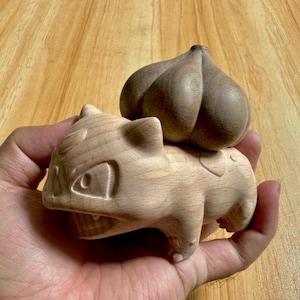 Puede incluir: Una pequeña figura de madera tallada a mano de una criatura con un cuerpo liso y claro y una estructura oscura, bulbosa y segmentada en la espalda. La figura presenta orejas puntiagudas, ojos definidos y una boca ancha de madera.