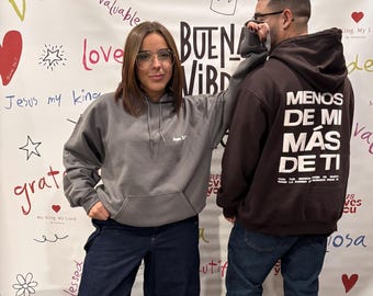 May include: Two people wearing gray and black hoodies with the text "BUENA VIBRA" and "MENOS DE MI MAS DE TI" on the wall behind them.