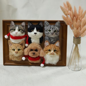 May include: A wooden frame with six felted animal heads, including cats and a dog, wearing red scarves with white pom-poms. The frame is on a white surface with a vase of dried flowers.
