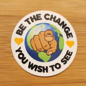 May include: A round sticker with the text "BE THE CHANGE YOU WISH TO SEE". The design features a pointing hand over a globe, with two yellow hearts. The sticker is white with black and colored graphics.