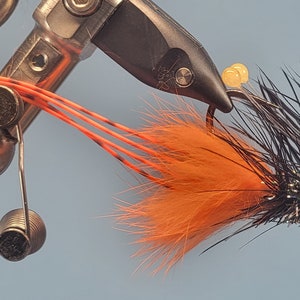 May include: A black and orange fly fishing lure with a red eye, tied on a hook and secured in a vise.