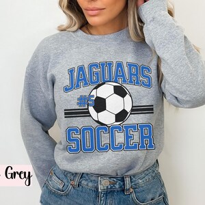 K&ouml;nnte beinhalten: Sportgraues Sweatshirt mit dem Text "JAGUARS SOCCER" in blauen Buchstaben und einer Fu&szlig;ballgrafik. Die Nummer "5" befindet sich &uuml;ber dem Fu&szlig;ball. Das Sweatshirt hat einen Rundhalsausschnitt.